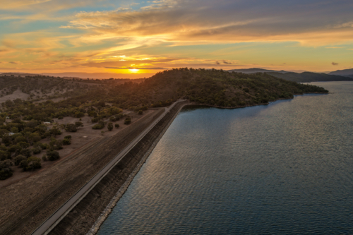 Aerial view of the Glenbawn Dam wall leading towards a sunset over the surrounding mountain range - Australian Stock Image