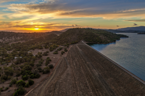 Aerial view of the Glenbawn Dam wall leading towards a sunset over the surrounding mountain range - Australian Stock Image
