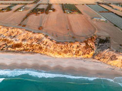 Aerial view of waves on a sandy beach below barren cliffs and narrow farm lots with homes - Australian Stock Image