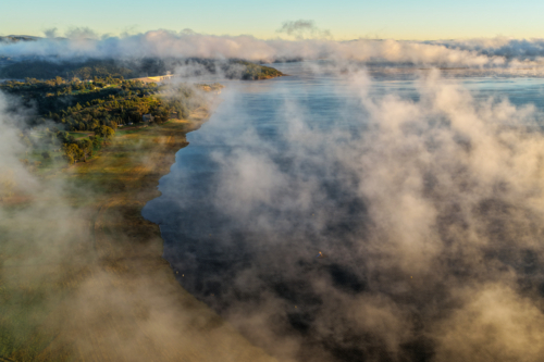 Aerial view over Lake Keepit Dam and the nearby campgrounds as fog drifts across the lake. - Australian Stock Image