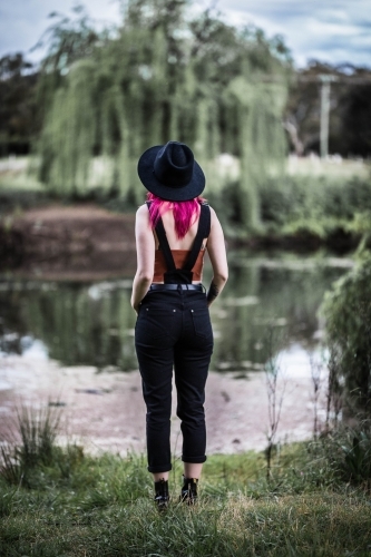 Alternative woman with pink hair and hat in overalls standing looking over river water