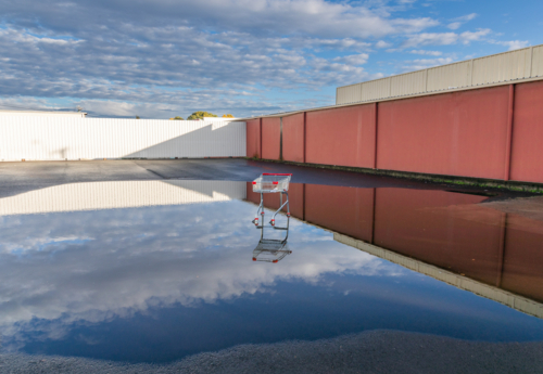 An abandoned shopping trolley in a puddle reflecting the sky and a long  fence - Australian Stock Image
