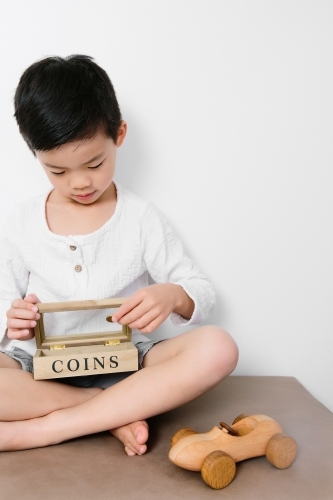 An asian child saving his coins in his money box with a wooden car toy beside him