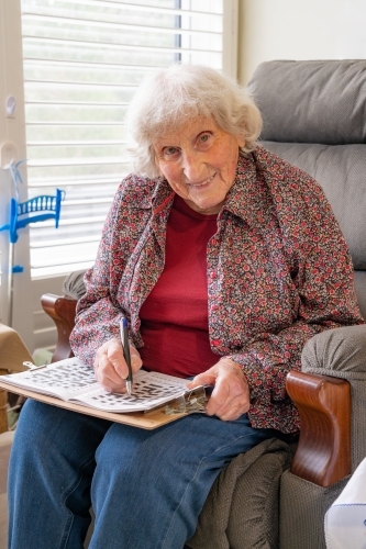 An elderly lady sitting in a chair doing a crossword and smiling - Australian Stock Image