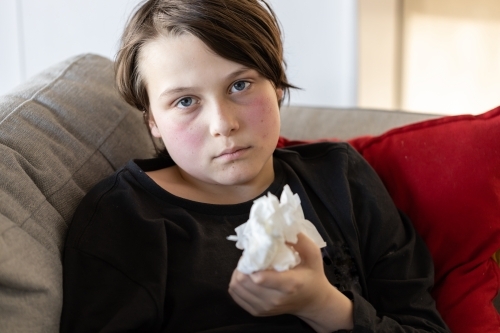 An unwell sick preteen boy lying on a couch holding some used tissues - Australian Stock Image
