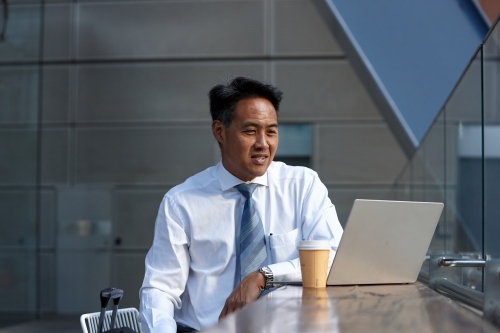 Asian businessman having a coffee whilst using laptop - Australian Stock Image