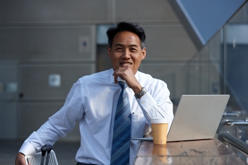 Asian businessman having a coffee whilst using laptop - Australian Stock Image