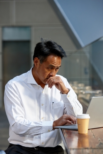 Asian businessman having a coffee whilst using laptop - Australian Stock Image