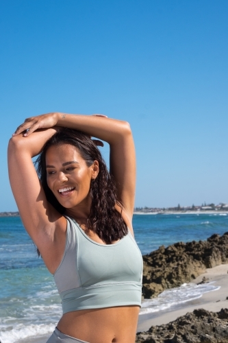Athletic woman in gym clothing stretching arms over head on coastal background - Australian Stock Image