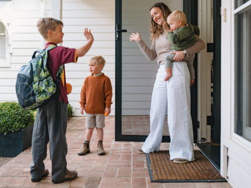 Aussie mum carrying toddler boy waving goodbye to older son leaving for school at the porch - Australian Stock Image