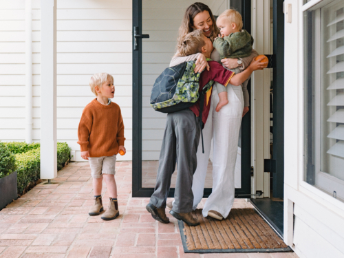 Aussie mum carrying toddler boy while hugging and kissing older son leaving for school at the porch - Australian Stock Image