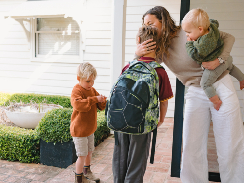 Aussie mum carrying toddler boy while hugging and kissing older son leaving for school at the porch - Australian Stock Image