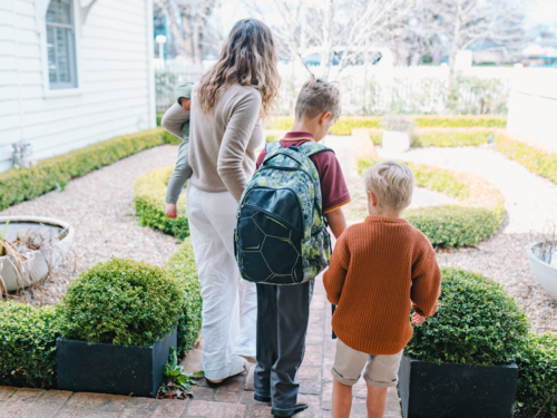 Aussie mum walking son with backpack on front porch while carrying toddler son on hip - Australian Stock Image