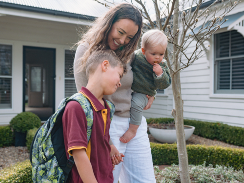 Aussie mum walking with young son while carrying her toddler on her hip to do school drop-off - Australian Stock Image