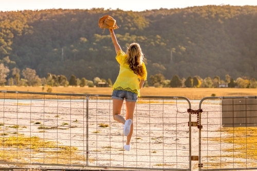 Aussie woman standing on a rural farm gate with one arm raised holding hat - Australian Stock Image
