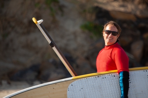 Australian surfer carrying Stand Up Paddle board on the beach - Australian Stock Image