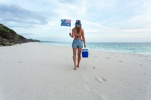 Australian woman in bikini and denim shorts walking along a beach with esky