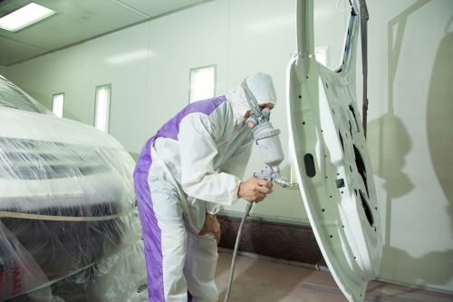 Auto painter in protective suit and mask, spraying a car in painting booth - Australian Stock Image