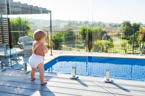 Baby standing at glass pool fence looking at water safe from drowning - Australian Stock Image