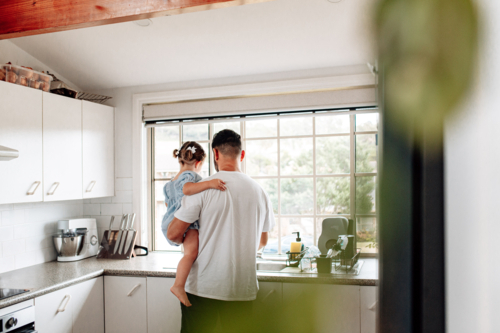 Back view of father carrying toddler daughter in the kitchen. - Australian Stock Image