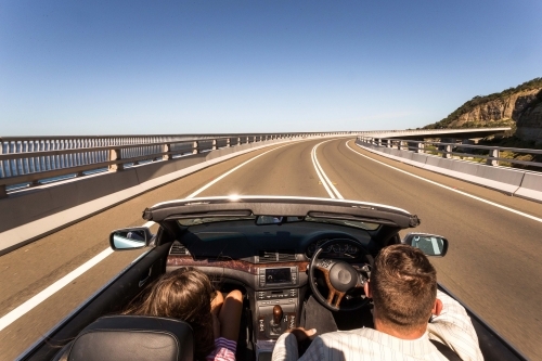 Back view shot of a man and woman sitting in a convertible car driving on the road near coast - Australian Stock Image