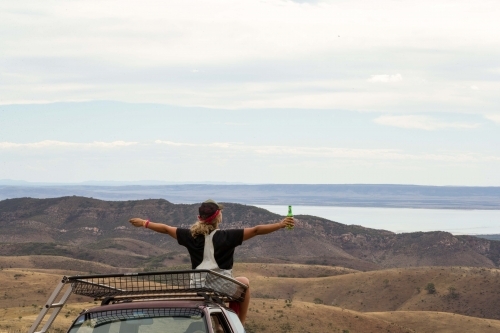 Back view shot of a woman holding a green bottle sitting on top of the car looking at the mountain - Australian Stock Image
