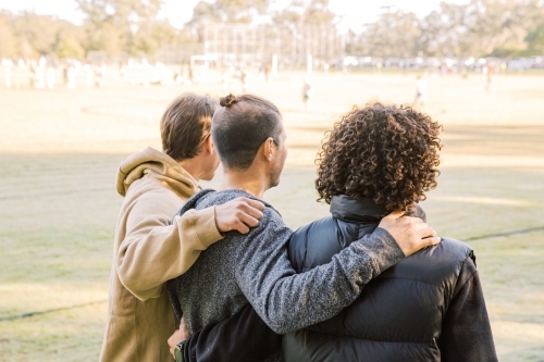 back view shot of three men one curly hair and one with a manbun with arms over each other's back - Australian Stock Image