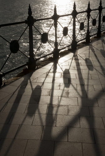 Backlit heritage safety fence throwing geometric shadows on paving with sunlit water behind