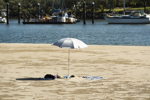 Beach umbrella in beach each scene at rivers edge with boats moored in water - Australian Stock Image