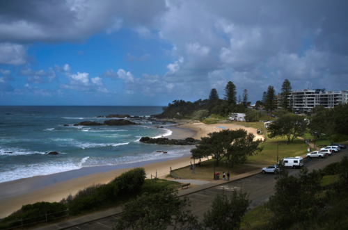 Beach with trees and buildings, sun and clouds - Australian Stock Image