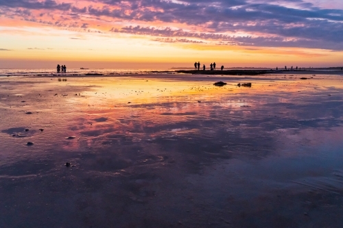 Beachcombers wandering in front of a vivid sunset sky and reflections - Australian Stock Image