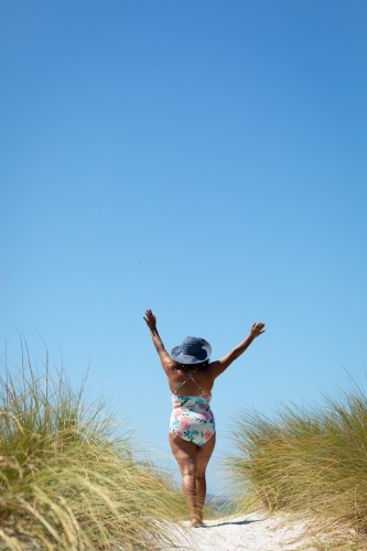 Big blue sky and woman in swimming costume walking towards the beach with arms raised - Australian Stock Image