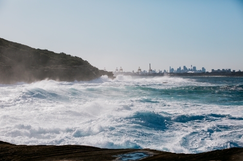 Big ocean swells along coast with cityscape in background - Australian Stock Image