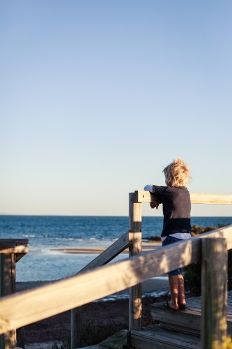 blonde boy gazing out over the ocean from wooden stairs to beach