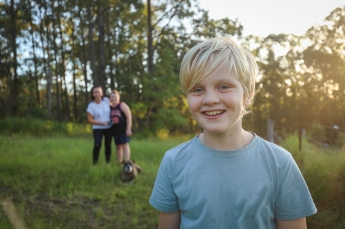 Blonde Caucasian boy smiling in the bush with mother and brother hugging out of focus - Australian Stock Image