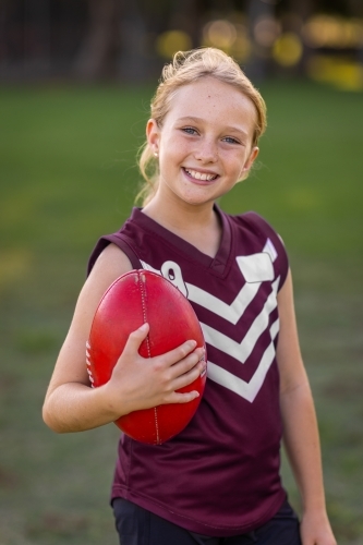 blonde girl holding red leather football and wearing football uniform - Australian Stock Image