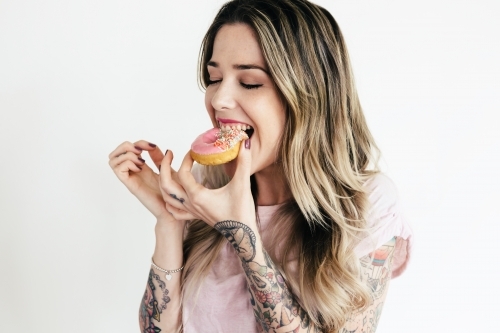Blonde girl in a pink t shirt biting into a pink iced donut - Australian Stock Image