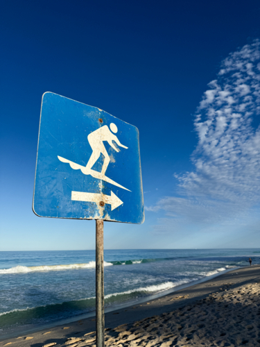 Blue Beach sign directing surfers in morning light with small waves and coastline - Australian Stock Image