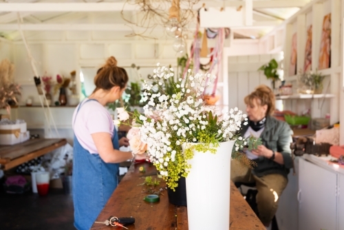Blurred female florist and assistant at work bench with vase of baby's breath flowers in foreground - Australian Stock Image