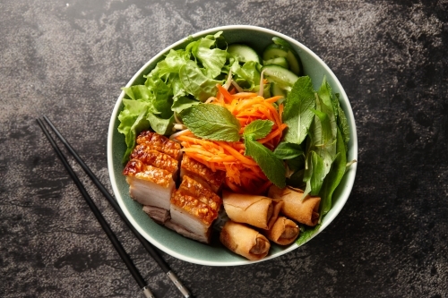 bowl greens with fried pork, spring rolls and carrots with black chopsticks on the side - Australian Stock Image