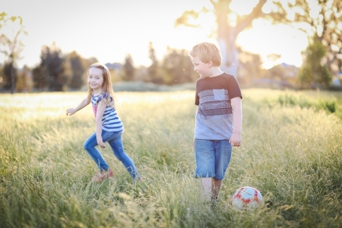 Boy and girl running through long grass playing soccer at the park. Aussie kids having fun outdoors - Australian Stock Image