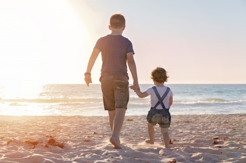 Boy And Little Girl Walking Towards The Ocean At Golden Sunset