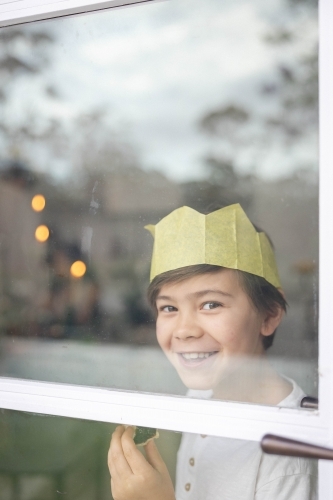 Boy at window wearing a Christmas crown and eating a mince pie - Australian Stock Image