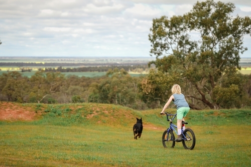 Boy bike riding with rottweiler dog on grass with view of farmland in the distance - Australian Stock Image