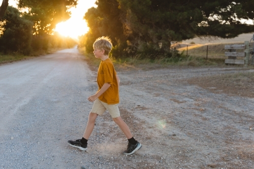 Boy crossing gravel country road at sunset wearing an orange top - Australian Stock Image