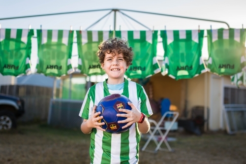 Boy holding soccer ball with team shirts hanging behind - Australian Stock Image
