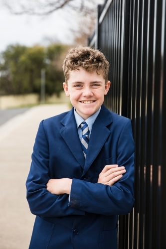 Boy in school uniform smiling with arms crossed leaning against fence at front of school - Australian Stock Image