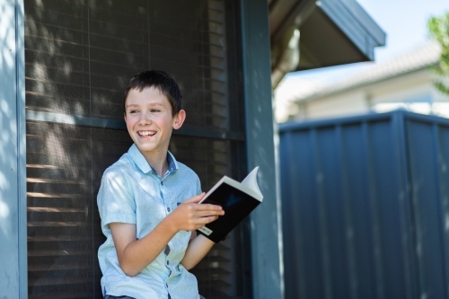 Boy laughs as he reads book outside next to house window - Australian Stock Image
