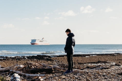 Boy looking at driftwood on beach with cruise ship in background