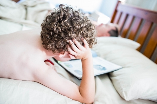 Boy lying on stomach on bed reading book with head propped on hand